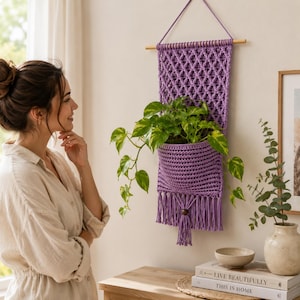 May include: A purple macrame wall hanging with a diamond-patterned top section and a solid pocket holding a green pothos plant. The piece is suspended from a wooden dowel with a string and features a bottom fringe with a single wooden bead.