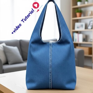 Op de afbeelding: Een blauwe denim tote bag met een enkele schouderband. De tas heeft een verticale naad in het midden en staat op een houten tafel. De woorden "+video Tutorial" staan in de linkerbovenhoek.