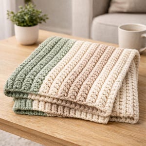 May include: A folded, striped crochet blanket in shades of green, beige, and tan. The blanket is made of a soft, textured yarn and is folded neatly on a wooden table. The crochet pattern creates a ribbed effect.