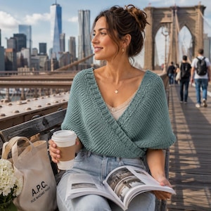 May include: A teal knitted wrap-style top with a V-neckline and ribbed trim, worn over a light-colored camisole. The garment features a textured knit pattern and short sleeves, paired with light blue denim jeans and a beige canvas tote bag.
