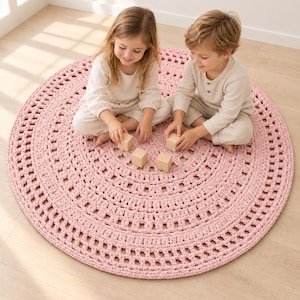 May include: A large, circular, light pink crochet rug featuring a textured, open-work pattern with concentric rings. The rug is crafted from thick yarn and displays a repeating geometric design throughout its surface, providing a soft, round base.