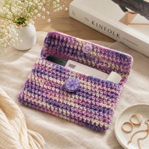 May include: A crocheted clutch bag in a mix of purple, lavender, and cream yarn. The open bag reveals a phone and other items. A flower-shaped button secures the flap. The clutch is on a neutral surface, with a book and vase in the background.