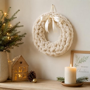 May include: A cream-colored, chunky knit wreath with a satin bow hangs on a wall. The wreath is a festive decoration. A small wooden house with a star cutout and a lit candle are on a wooden surface below the wreath.