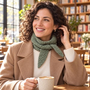 Peut inclure: Une écharpe triangulaire en tricot vert sauge avec un motif texturé, portée autour du cou sur un pull côtelé crème et un manteau en laine beige. L'écharpe présente une extrémité pointue et une construction en laine épaisse.
