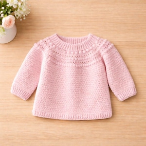 Puede incluir: Un suéter rosa claro para bebé con mangas largas. El suéter presenta un patrón de punto texturizado y un cuello redondo con detalles de bolitas. La prenda está hecha de hilo suave, adecuada para bebés y niños pequeños.
