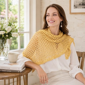 May include: A golden yellow crocheted shawl with a diamond pattern. The shawl is draped over the shoulders. The person is sitting at a wooden table with a cup and books. A vase of white flowers is in the background.