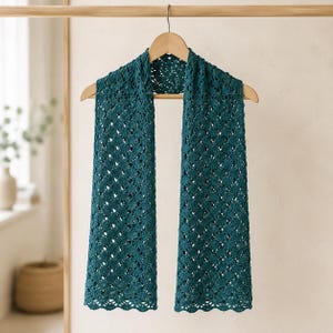 Può includere: Una sciarpa color verde ottanio scuro caratterizzata da un intricato motivo a uncinetto traforato con un design a rombi ripetuto. La sciarpa è drappeggiata su una gruccia di legno, mettendo in risalto la trama del filato e i bordi smerlati.
