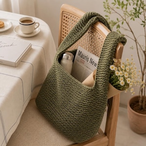 Könnte beinhalten: Eine olivgrüne Häkeltasche im Hobo-Stil mit einem dicken Schulterriemen. Die Tasche enthält eine silberne Metallwasserflasche, eine Zeitung mit dem sichtbaren Text Mauris News und einen kleinen Strauß weißer Gänseblümchen in braunem Papier.