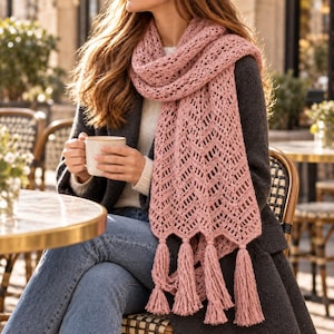 Puede incluir: Una bufanda de ganchillo en color rosa empolvado con un intrincado patrón de chevrón y largas borlas gruesas en los extremos. La bufanda está colocada alrededor del cuello, mostrando la textura calada del hilo y el detalle de flecos anudados.