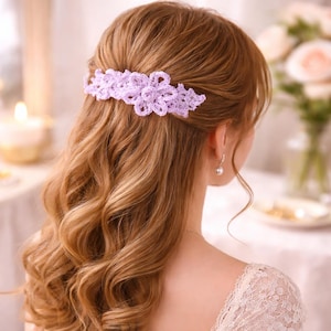 Puede incluir: Horquilla de pelo de ganchillo lila claro con diseño floral. La horquilla se coloca en cabello largo y ondulado, de color castaño claro. El fondo está borroso, con una vela y flores. Un accesorio decorativo.