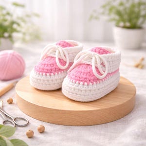 May include: Hand-knitted baby booties, designed to resemble trainers. The shoes are white with pink detailing and white laces. The booties are made from a soft, knitted material. A ball of pink yarn and scissors are in the background.