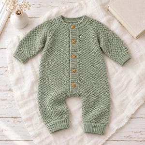 May include: A sage green knitted romper featuring a textured diamond pattern, long sleeves, and a ribbed crew neckline. The front placket is secured with five small, light brown wooden buttons shaped like flowers, and the cuffs are ribbed.