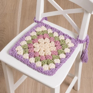 May include: A square, crocheted chair cushion in a granny square pattern. The cushion features a central cream-colored square surrounded by pink, green, and cream squares, all bordered by a lavender edge. It has lavender ties with a tassel, and is placed on a white chair.