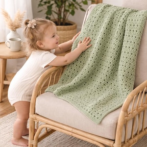 May include: A light green crochet blanket draped over a wicker chair. The blanket has a textured, open weave design. The chair is made of light brown wicker and has a neutral-colored cushion. A young child is touching the blanket.