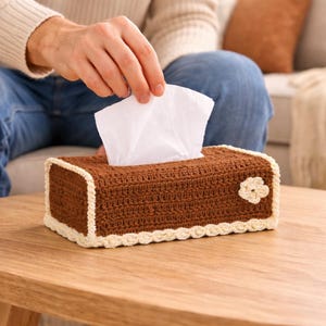 May include: A brown crochet tissue box with a white flower accent. The box has a cream-colored trim and is sitting on a wooden table. White tissues are pulled from the top of the box.