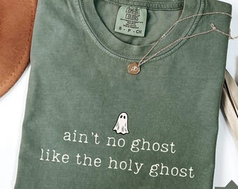 以下が含まれることがあります： 小さな白い幽霊のグラフィックと「ain't no ghost like the holy ghost」という白い文字がプリントされた、モスグリーンの半袖Tシャツ。クルーネックで、襟の内側にはComfort Colorsのブランドラベルが付いています。