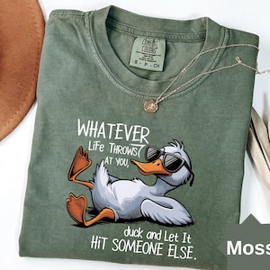 May include: A moss green t-shirt with a graphic of a duck wearing sunglasses and the text "WHATEVER Life THROWS AT YOU, duck and Let It HIT SOMEONE ELSE." A gold necklace is draped across the shirt.