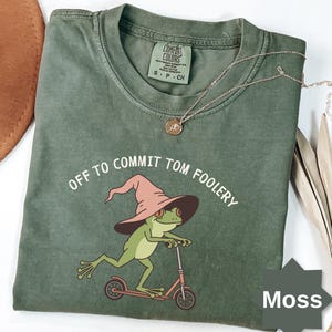 May include: Moss green Comfort Colors t-shirt with the phrase "OFF TO COMMIT TOM FOOLERY" and a cartoon frog wearing a witch hat and riding a scooter. A gold necklace is draped across the shirt.