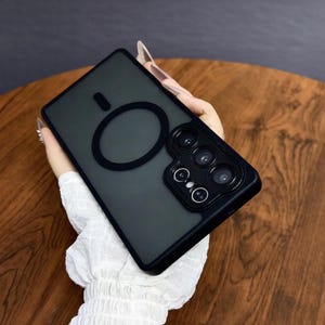 May include: Black phone case with a matte finish and a circular design element. The case has a raised section for the camera lenses. The case is held against a wooden surface.