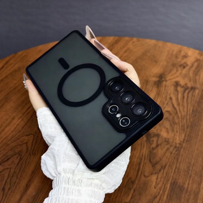 May include: Black phone case with a matte finish and a circular design element. The case has a raised section for the camera lenses. The case is held against a wooden surface.