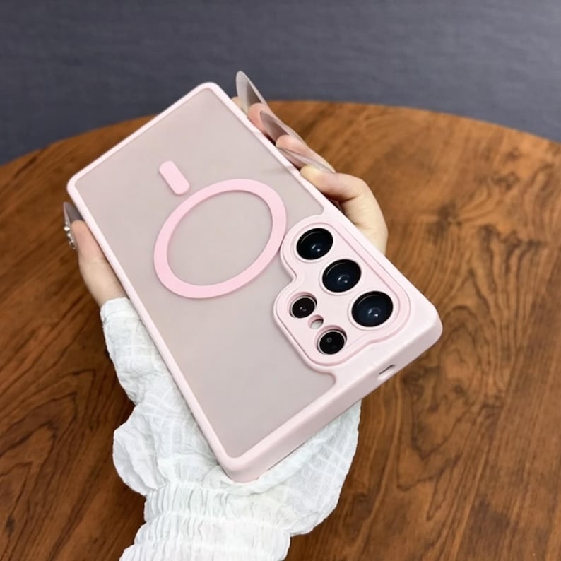 May include: A light pink phone case with a matching pink circular design and camera module. The case has a clear back and a pink border. The phone case is designed to protect the phone from damage.
