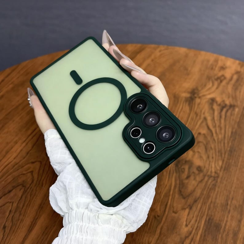 May include: A sage green phone case with a dark green border and camera module. The case features a circular design on the back. The camera module has three lenses and two smaller circles. The case is held by a person.