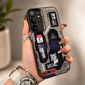 Peut inclure: Une coque de smartphone grise au design mécanique avec des accents noirs, du texte incluant "WARNING", "STAGE 2+", "HACKED" et "LOADING", ainsi qu'un code QR, une icône d'éclair jaune et divers labels techniques et motifs géométriques.