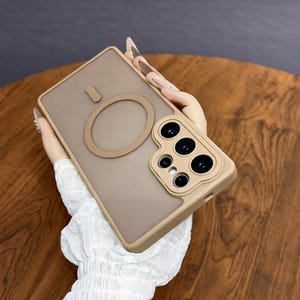 May include: A tan phone case with a circular magnetic attachment and a camera module. The case has a matte finish and a raised edge around the camera. The case is held in a person's hand.