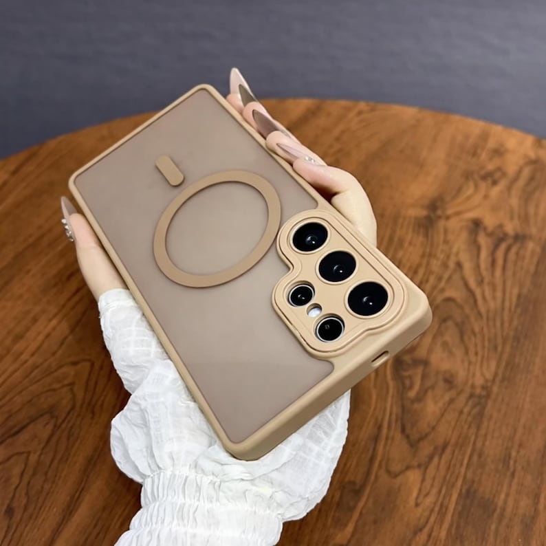 May include: A tan phone case with a circular magnetic attachment and a camera module. The case has a matte finish and a raised edge around the camera. The case is held in a person's hand.