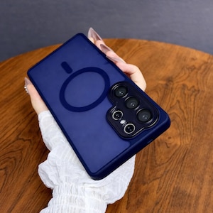 May include: A navy blue phone case with a circular design and a camera module. The case has a matte finish and is designed to protect the phone. The case is shown on a wooden surface.