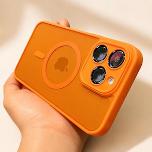 May include: A bright orange smartphone case featuring a circular magnetic charging ring and a central Apple logo. The case includes a raised square camera module housing three circular lenses and a small circular flash, all in a matching orange tone.