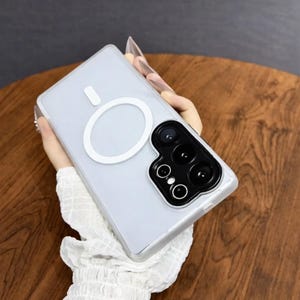 May include: A clear phone case with a white magnetic ring and camera module. The case is held by a person with long nails. The phone case is designed to protect the phone from scratches and damage.