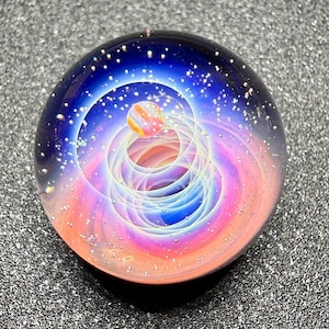 May include: A glass paperweight with a celestial design. The sphere features a swirling galaxy of pink, blue, and orange hues, with a small planet at the center. The outer layer is a deep blue, speckled with white dots.