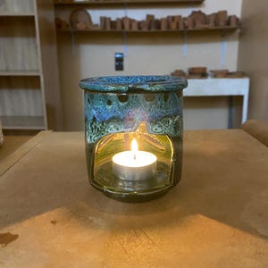 Op de afbeelding: Een keramische etherische olie diffuser met een blauwgroen glazuur. Een brandende waxinelichtkaars zit in de diffuser, zichtbaar door een decoratieve opening. De diffuser staat op een bruin oppervlak.