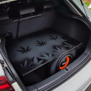 Puede incluir: Un recinto de subwoofer negro con un diseño de hoja de cannabis está instalado en el maletero de un coche blanco. El subwoofer tiene un anillo naranja alrededor del altavoz.