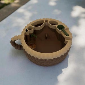 Może przedstawiać: Brązowy i beżowy miniaturowy ogród w kształcie filiżanki do herbaty. Zawiera małe zielone narzędzia ogrodnicze, grosz i centralny brązowy obszar. Filiżanka ma teksturowany, koszopodobny wzór wokół krawędzi i uchwytu.