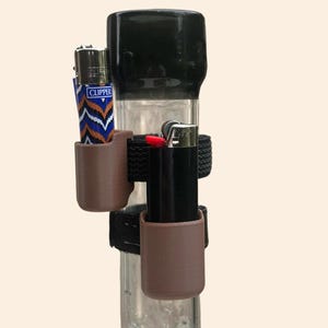 以下が含まれることがあります： 黒いマウスピースと、茶色のホルダーと黒いストラップで固定された2つのライターが付いた、透明なガラス製の水パイプ。1つのライターは青と白のシェブロン柄で、もう1つは黒です。