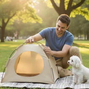 May include: A beige pet tent with a circular opening is set up on a checkered blanket in a park. A small white dog sits nearby, and a person is setting up the tent. The scene is bathed in sunlight.