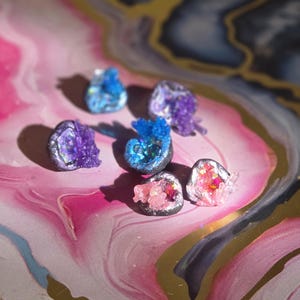 May include: A collection of six small, irregular geode-style ornaments featuring metallic silver outer shells and vibrant, crystalline interiors in shades of blue, purple, and pink, with sparkling glitter accents embedded within each piece.