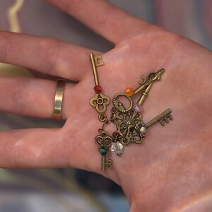May include: A collection of antique-style brass keys attached to a central ring, featuring intricate filigree designs and small, colorful faceted beads in red, orange, clear, and green hues, creating a decorative set of metal skeleton keys.