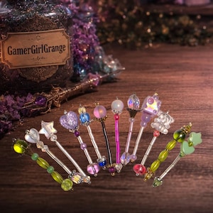 May include: An assortment of decorative wands with colourful crystal and bead designs. Each wand has a unique top, such as stars, hearts, and spheres. The wands are displayed on a wooden surface, with a jar labelled "GamerGirlGrange" in the background.