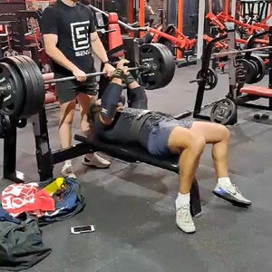 Puede incluir: Una persona está haciendo press de banca en un gimnasio. La persona está acostada en un banco negro y levantando una barra con pesas. Otra persona está de pie cerca, ayudando. El gimnasio tiene equipos rojos y negros.