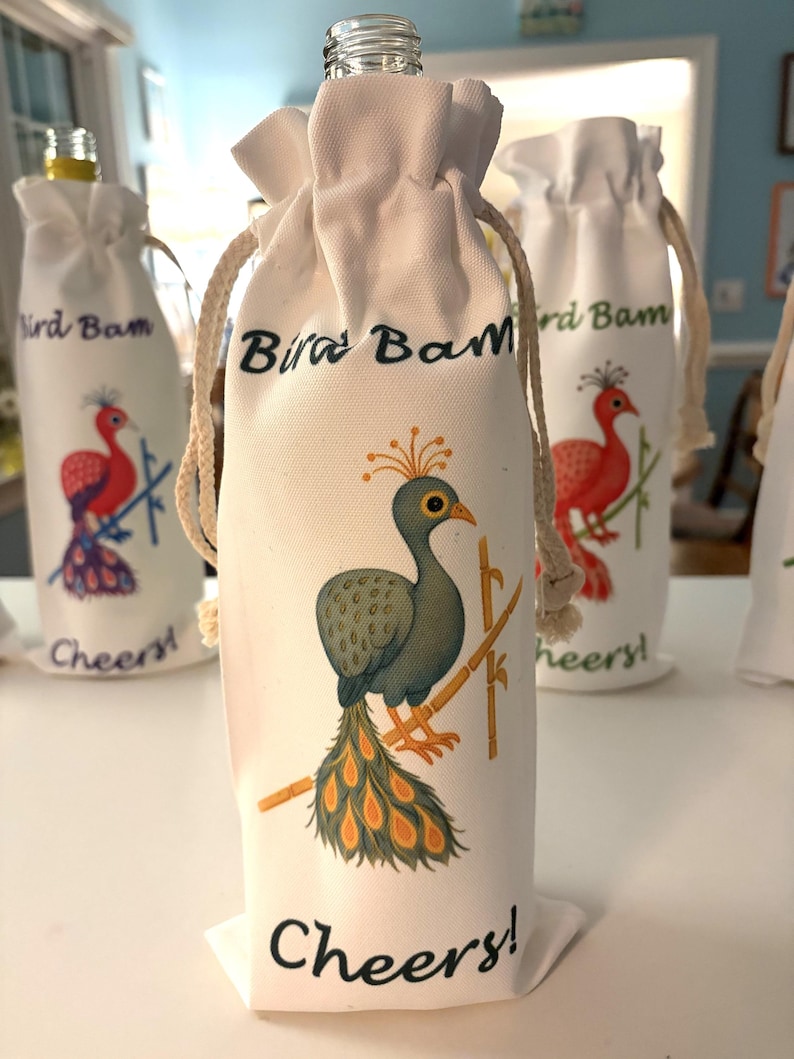 May include: White fabric wine bottle bags with drawstring closures. Each bag features a colorful illustration of a bird and the text "Bird Bam" and "Cheers!". The bird illustrations are in shades of red, green, and yellow.
