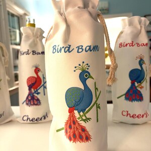 May include: White fabric wine bottle bags with a drawstring closure. Each bag features a colorful peacock illustration with the text "Bird Bam" and "Cheers!". The peacock has blue, red, and green accents.