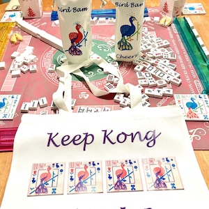 May include: A Mahjong game setup with tiles, two white bottle bags with peacock designs and the text "Bird Bam" and "Bird Ban". A white tote bag with the text "Keep Kong ...and Mah Jong" is in the foreground.