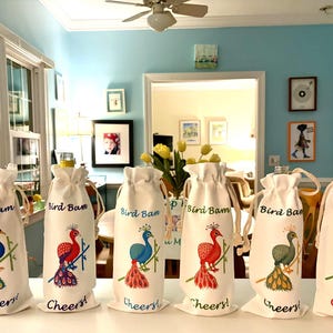 Puede incluir: Bolsas blancas para botellas de vino con cierres de cordón. Cada bolsa presenta una colorida ilustración de pavo real y las palabras "Bird Bam" y "Cheers!". Los diseños de pavo real varían en color, incluyendo azul, rojo y verde.