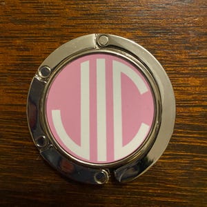 May include: A silver-colored, circular purse hook with a pink center. The center features the white initials "JIC". The hook is designed to hang from a table's edge, providing a convenient place to store a purse or handbag.