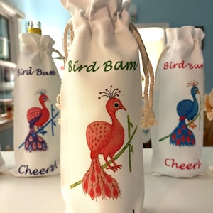 May include: White wine bottle bags with peacock designs in red, blue, and pink. The text "Bird Bam" and "Cheers!" are printed on the bags. The bags have a natural rope drawstring closure.