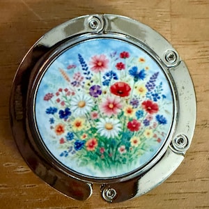 May include: A silver-toned purse hook with a round, floral design. The center features a watercolor painting of colorful wildflowers, including daisies, poppies, and lavender, against a light blue background. The hook is designed to hang from a table.