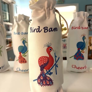 May include: White fabric wine bottle bags with a drawstring closure. Each bag features a colourful peacock illustration and the words "Bird Bam" and "Cheers!". A yellow bottle is visible in the top of one bag.